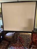 Front view of the projector screen fully extended showing the white projection surface and tripod stand on carpet.