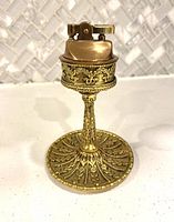 Vintage brass table lighter with ornate detailed brass pedestal base photographed on a white surface with a tiled backsplash.