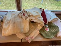 Whole pile of assorted table linens including tablecloths, placemats, and runners seen stacked on a sill, showing the variety of fabrics and colors.