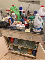 Cleaning supplies including detergent, sprays, brushes, clothespins, gloves, and various bottles on a black countertop cabinet with open doors showing additional supplies on shelves beneath.