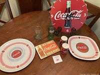 Photo showing Coca-Cola branded salt and pepper shakers, two plates with checkered borders, a glass bottle, a glass tumbler, a metal plaque, and a red wall clock with Coca-Cola logo and bottle graphic.