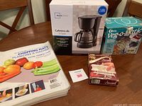 Mainstays coffee maker box, Ottter Veg-o-Matic box, flexible chopping mats, and meat tenderizer box on a table.