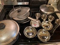 Dutch oven, paella pan, saucepan, stainless steel cups, rice bowls, and spatter guards arranged on a stovetop.