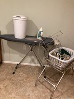 Photo showing ironing board with iron and trash can on top, vintage wire laundry cart with laundry basket filled with hangers nearby