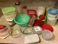 Overview of assorted kitchen storage containers, glass bowls, plastic containers with various lids including red and blue, green plastic colander, plastic bottles