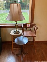 Full view of the lamp with raw silk shade on a square wooden stool, round small table plant stand, and wooden high chair shown together near a window.