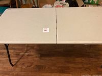 Photo of the full rectangular beige folding table top with metal legs open and set up on wooden floor.