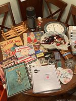 Wide shot of lot items on wooden table showing vintage magazines, monkey fist keychains, thermos, clock, ashtrays, shoelaces, and other items.