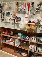 Photo showing kitchen wall pegboard with hanging utensils, lightbulbs, electrical tools, and collectibles, along with shelving containing tins, containers, and kitchen goods.