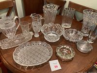 Photo showing a collection of assorted crystal and cut glass items including pitchers, vases, bowls, and serving dishes arranged on a wooden table.