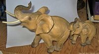 Two porcelain elephant figurines, adult and calf, shown on wood surface with background objects, highlighting size and condition.