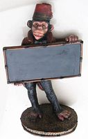 Front view of the monkey statue holding blank message board with red fez hat and brown jacket, standing on textured oval base.