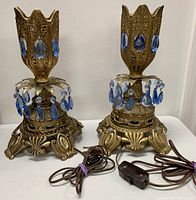 Pair of gothic brass table lamps with blue prism crystal drops on a white surface, showing ornate bases and cords with switches.