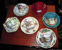Top view of six different vintage English porcelain teacups on matching saucers arranged on a wooden table displaying various floral and scenic patterns with some gold trim