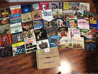 Photo showing 44 vinyl LP record covers laid out on wooden floor with a cardboard box labeled 'This Side Up' in front. The covers display a variety of music styles and artists.