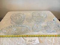 Five clear crystal glass pieces arranged on a table with white cloth and measuring tape; includes round bowls, divided tray, and lidded dish.