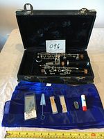 A velvet-lined black carrying case containing disassembled EVETTE clarinet parts with keys and cork joints visible, lying open on a table. Blue cloth holding accessories arranged in front of the case.