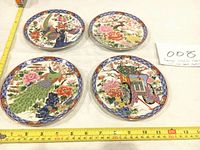 Four Japanese collectible fine porcelain plates arranged on a table with measurement tape showing approximately 6.5 inches diameter.
