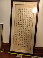 Full view of large framed Chinese calligraphy artwork showing text in vertical columns and artist stamps at bottom with artist signature. Wood frame and glass cover visible.