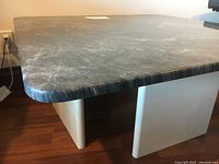 Angled side view showing rounded edges of dark polished marble top with white geometric base on wood floor.