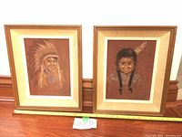 Two framed pastel drawings side by side depicting a First Nation boy and girl on felt, placed on floor with measuring tape.