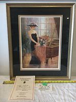 Photo of framed lithograph art "The Music Room" showing a woman in Victorian black dress with floral hat near a piano with flowers, with authentication certificate and measuring tape in front.