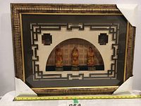 Front view of three small Chinese fortune statues framed and mounted in an ornate gold frame with geometric design and protective corner covers.