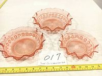Top-down view showing three pink pressed glass bowls with ruffled edges and patterned rims placed next to a measuring tape.
