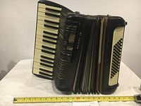 Full side angled view of vintage black Hohner VERDI IA piano accordion showing piano keys and bellows with measuring tape.