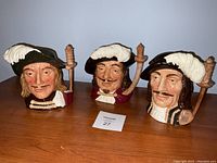 Three large Royal Doulton Toby jugs representing The Three Musketeers, showing all three grouped together on a wooden surface with clear visibility of facial and handle details.