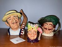 Photo showing all three Royal Doulton Toby jugs together: a man with a sailor hat and striped shirt, a man with a green hat and flamingo handle, and a man with green feathered hat and mustache.