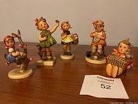 Five colorful Hummel figurines arranged on a wooden surface, including a girl on a tree branch, girls with flowers and basket, a knitting child, and a boy playing accordion.