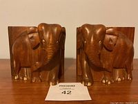 Front view of two matching carved wooden elephant bookends showing detailed elephant heads and front legs, with some toes missing.