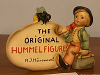 Front view of the Hummel retail display figurine showing a boy resting on a suitcase next to a sign stating 'The Original Hummel Figures M.J. Hummel' with a bee on top.