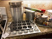Photo showing stack of aluminum bucket pots, 24-cup muffin tin, baking sheets, metal pots with lids, and a box of foil on kitchen countertop.