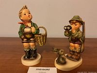 Photo of both Hummel figurines showing Village Boy with basket and Good Hunting boy with binoculars and rabbit.