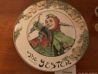 Full top-down view of the circular decorative plate showing the jester illustration, border foliage and fruit, and inscription 'The Jester'.
