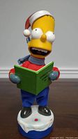 Front view of the Bart Simpson figurine showing it holding a green book and wearing a red Santa hat and blue scarf.