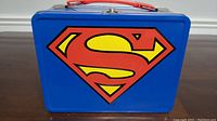 Front side of blue metal lunchbox with large yellow and red Superman 'S' logo.