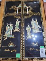 Front view of both black lacquered wall panels side by side showing intricate gold painted borders and traditional Chinese scenes with shell inlaid women figures.