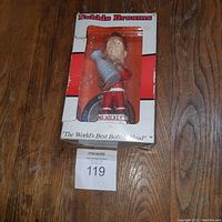 Front view of Gordie Howe bobble head in box showing figure dressed in red hockey gear holding a silver trophy with box branding visible.