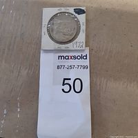 Top view of the coin holder showing the coin encased with a white cardboard holder and a MaxSold lot tag showing number 50.