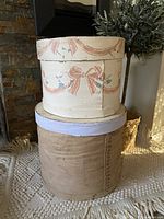 Two round wooden cheese boxes stacked, top box painted with pink ribbon design, bottom box plain natural wood.