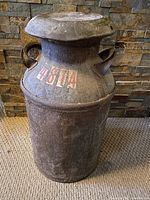 Antique metal milk can standing on carpet in front of stone wall, showing front view with visible rust and faded large white and red number decal '4814' near top.