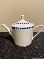 Side view of the white ceramic teapot with blue polka dots, showing the spout, handle and lid with knob.