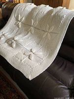 Cream hand knit afghan blanket draped over a dark leather sofa showing raised floral panel details and border texture.