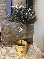 Full view of artificial olive tree standing in woven wicker basket with star accent, placed on a textured rug in front of a stone fireplace.