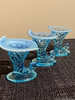 Three turquoise blue hobnail cornucopia shaped glass dishes with opalescent rims on black textured fabric surface, showing curling tail base and wide open mouth of each dish.