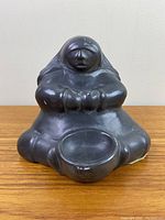Front view of the black soapstone Inuit figurine showing full seated figure with rounded form and bowl.