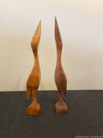 Pair of slim wooden crane figurines showing front view on black surface with plain wall background.